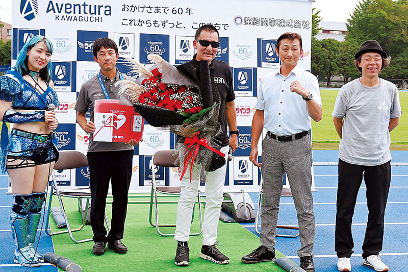 産経商事の藤田憲一本部長（右から2人目）からAEDの寄贈を受けたアヴェントゥーラ川口の長岡修理事長（同4人目）。中央は蝶野正洋さん＝川口市の青木町運動公園競技場