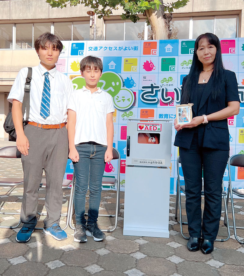 AEDの贈呈式に出席した（右から）桐田明日香さんの写真を手に持つ母親の寿子さん、弟の悠さん、真さん＝30日午後、埼玉県さいたま市北区の日進中学校