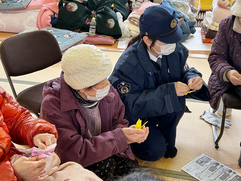 折り紙で鶴を折りながら被災者の話に耳を傾ける特別生活安全部隊の隊員（右）（県警提供）