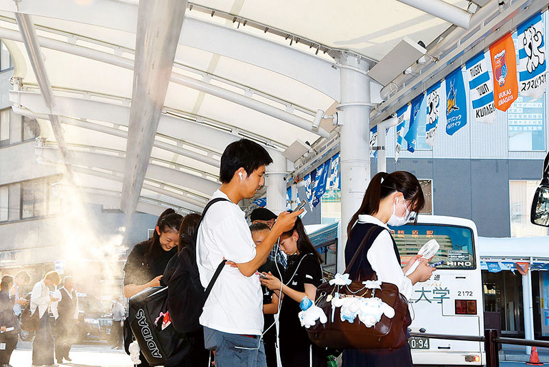 冷却ミストが作動したバス停で涼を取りながらバスの到着を待つ客＝14日午後4時45分ごろ、熊谷市筑波の熊谷駅北口