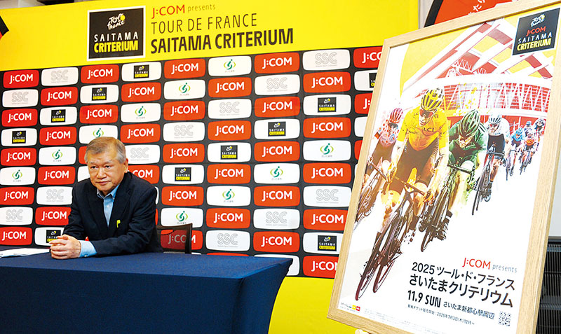 新たなメインビジュアルの脇で取材に応じる、さいたまスポーツコミッションの遠藤秀一会長＝1日、さいたま市浦和区のときわ会館