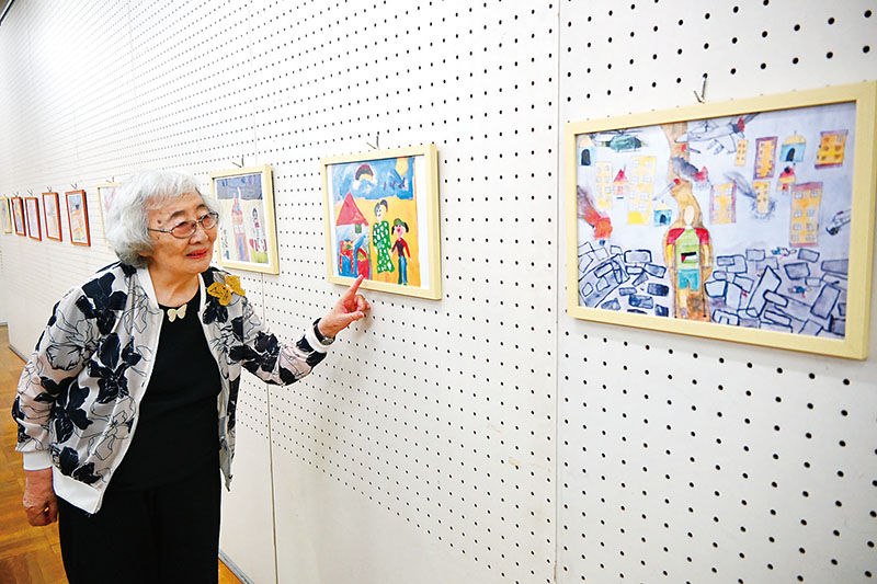 2023～24年ごろにガザの子どもたちが描いた絵を見る野村路子さん。右の絵はがれきの中、松葉づえを突いた人が歩く。爆弾が落とされ、建物が燃えている様子が描かれている＝3日、川越市内