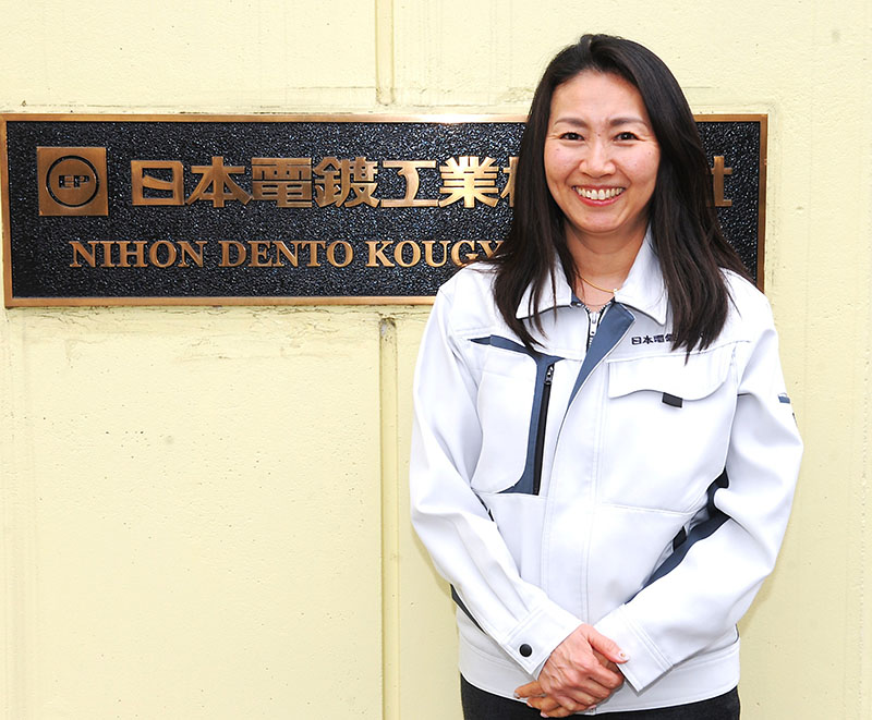 社名が刻まれた正門の前で笑顔を見せる日本電鍍工業の伊藤麻美社長