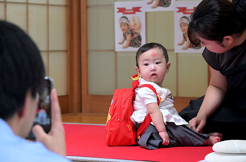 一升分の餅の入ったバッグを背負う赤ちゃんを撮影する親子