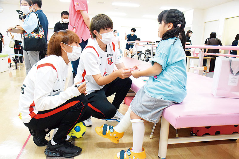 県立小児医療センターで子どもたちと交流する浦和レッズレディースの長嶋玲奈選手（中央）と丹野凜々香選手（左）＝さいたま市中央区