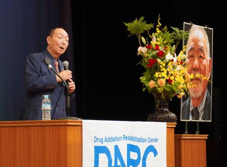 近藤恒夫さんの遺影が飾られた会場であいさつする、茨城ダルク代表の岩井喜代仁さん＝２７日午後、東京都台東区